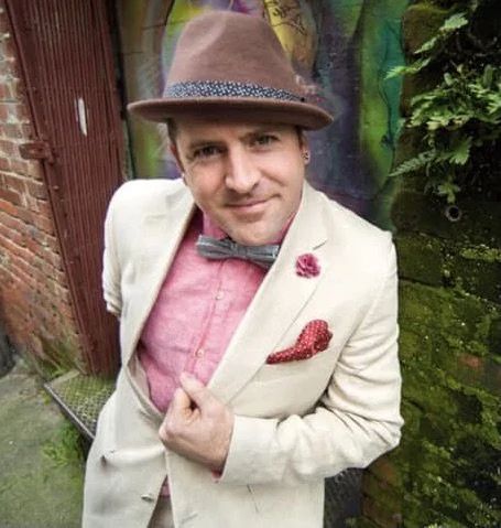 Bryce Williams in a cream jacket, pink shirt and fedora in front of a mural
